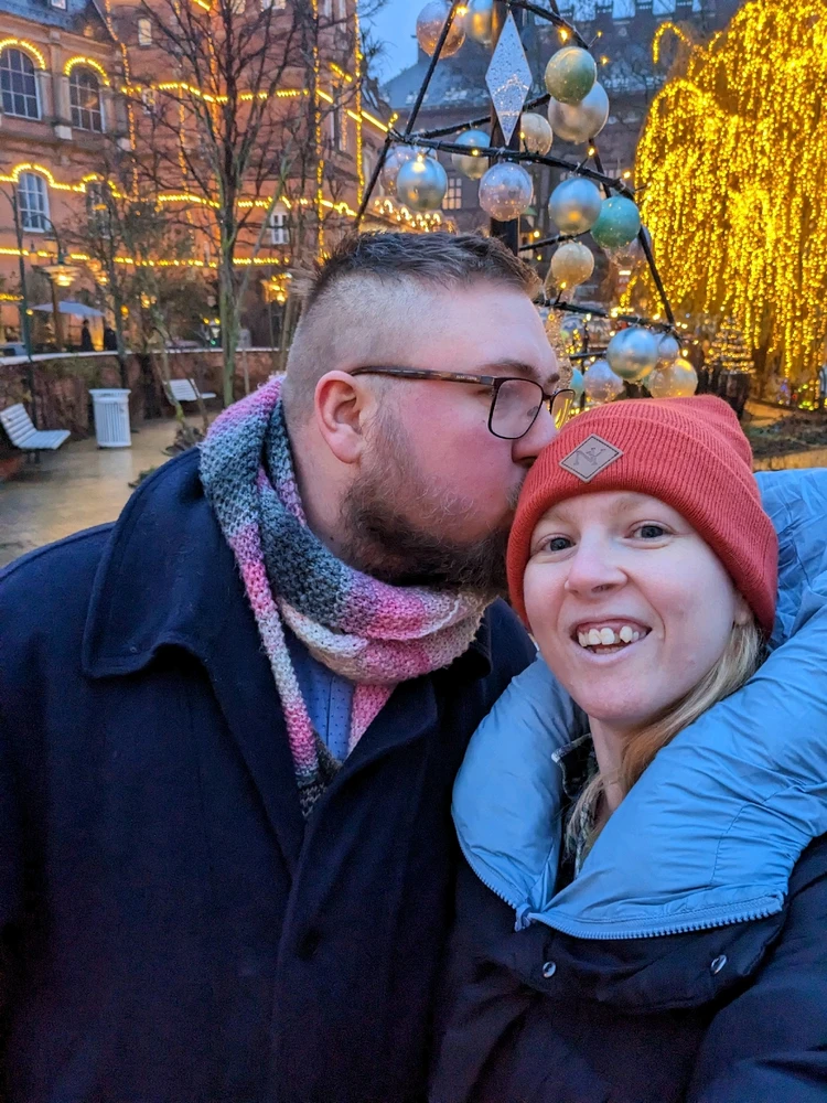 A couple in a winter setting. The man on the left is kissing the woman, on the right, on her head. She is smiling into camera