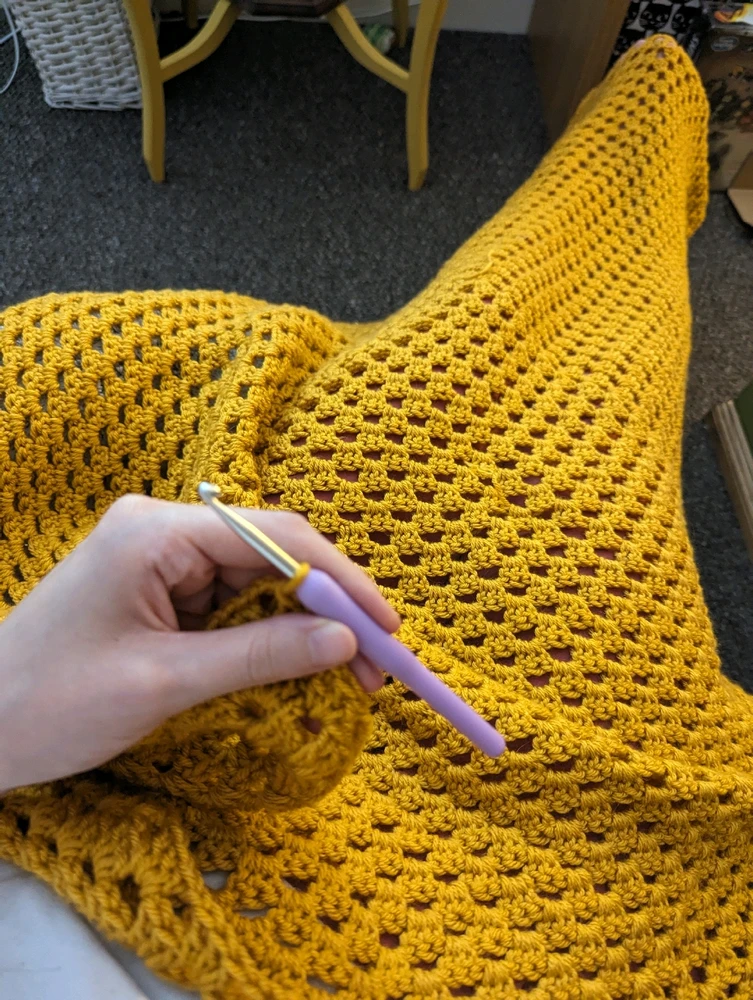 Photo of a granny square being crocheted in mustard yellow yarn. The square is big enough to cover Maisie's legs.