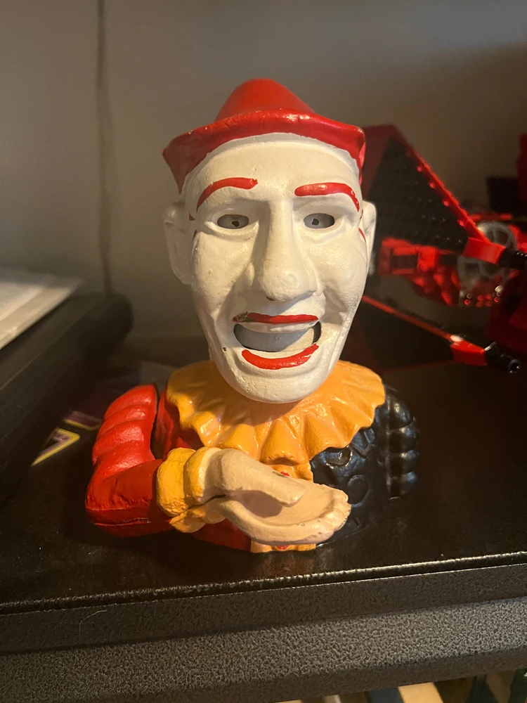 metal clown bank with red hat, eyebrows, mouth, and white face with moving eyes & mouth eats coin when lever is pulled.