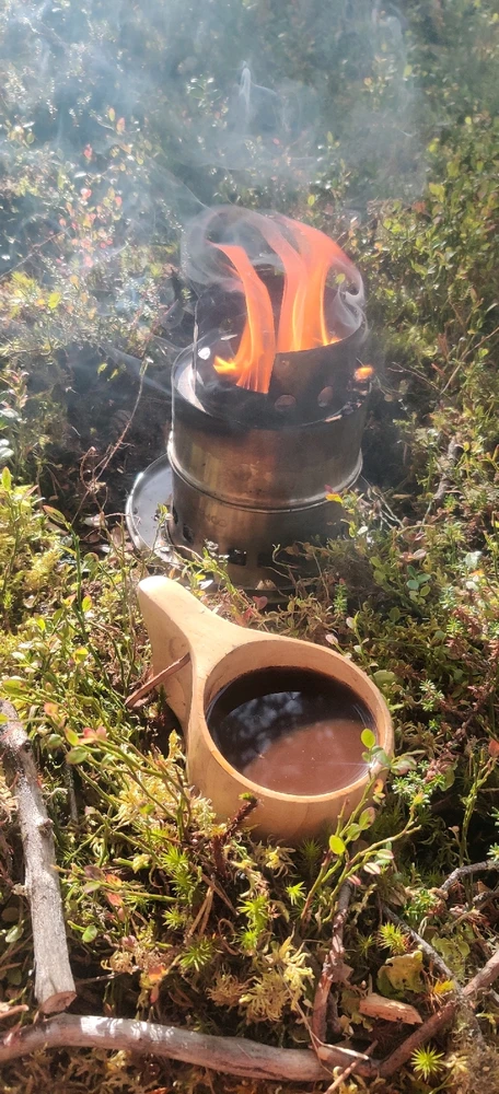 A kuksa cup filled with coffee sitting on a bed of moss with a twig stove on a protective plate behind it.