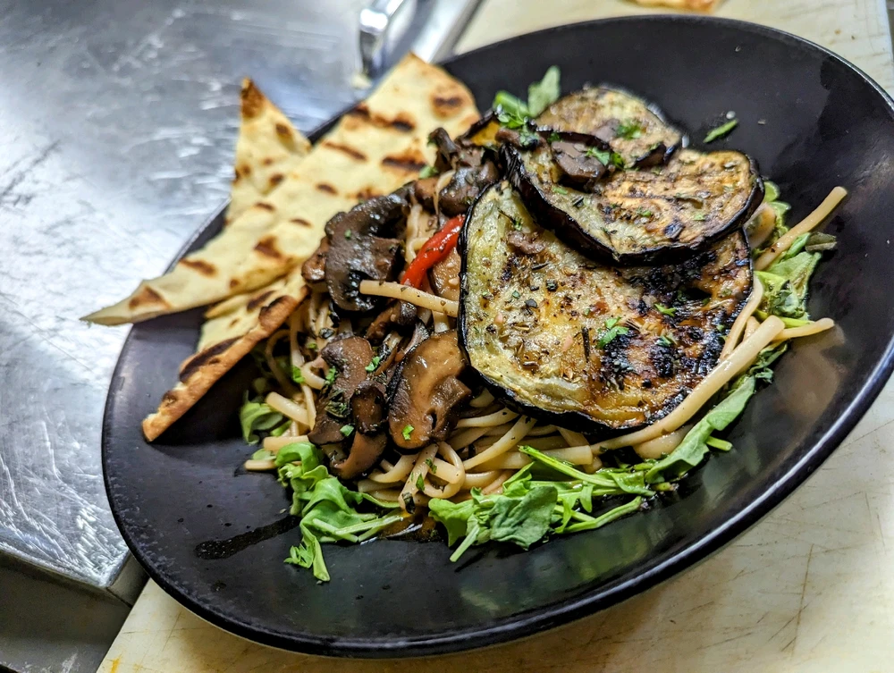 Black plate with linguine pasta tossed in a mushroom sauce, topped with grilled eggplant slices, and a side of grilled pita 