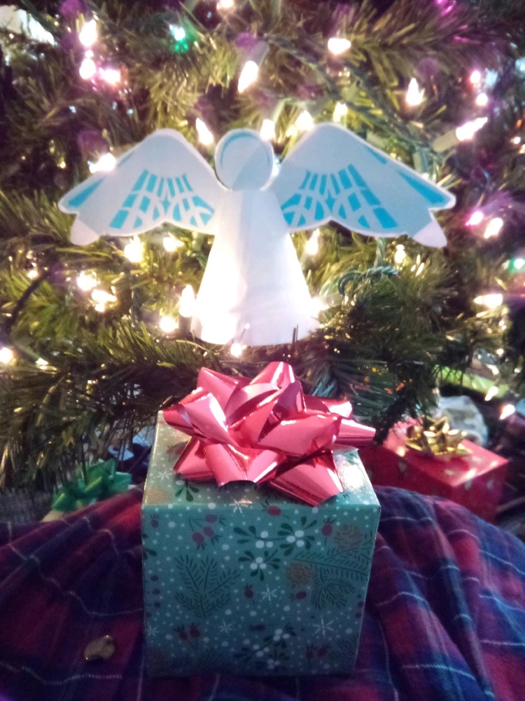 A paper angel sitting in a Christmas tree surrounded by lights. Below it is a green present with a bright red bow 
