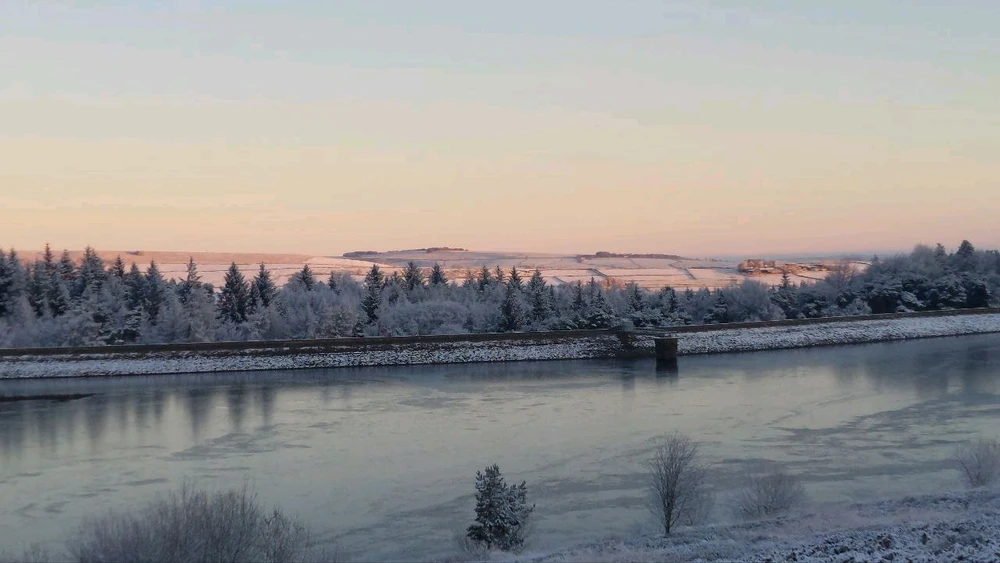 Dawn sun illuminating snowy landscape with a pink hue, lake in foreground.