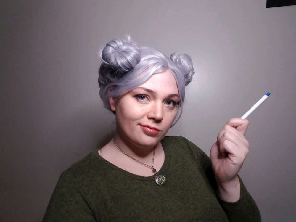A young-looking woman with a silver wig parted into two buns on top of her head, wearing a green sweater and a smirk. 