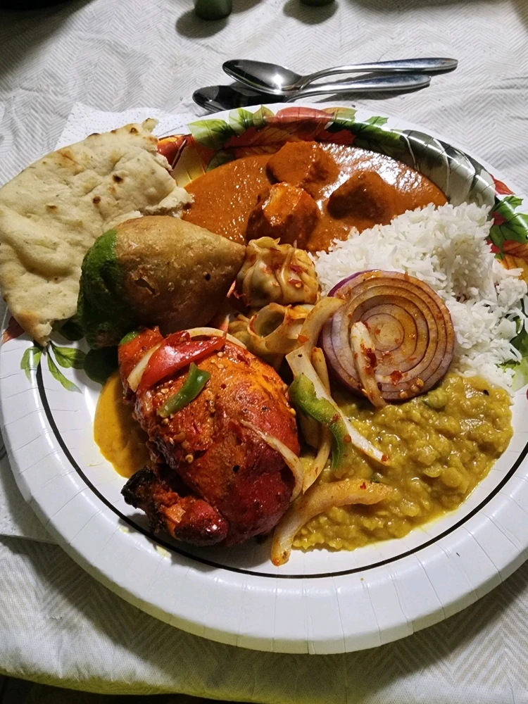 On this plate is Butter chicken, Vegetable Samosa, Garlic Naan, Spicy Tofu Momo, Jasmine Rice, Tarka Daal,Tandoori Chicken