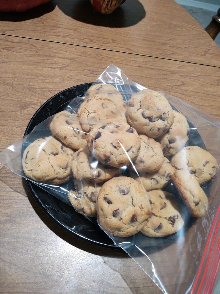 Brown table with a black plate on top with large fluffy chocolate chip cookies resting in a pyramid stack inside a zip lock