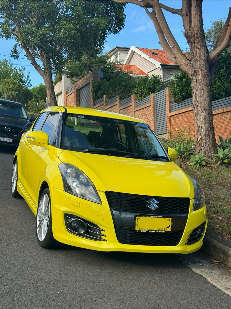 a photo of the front of a yellow suzuki swift sport parked at the side of the road on an incline. 
