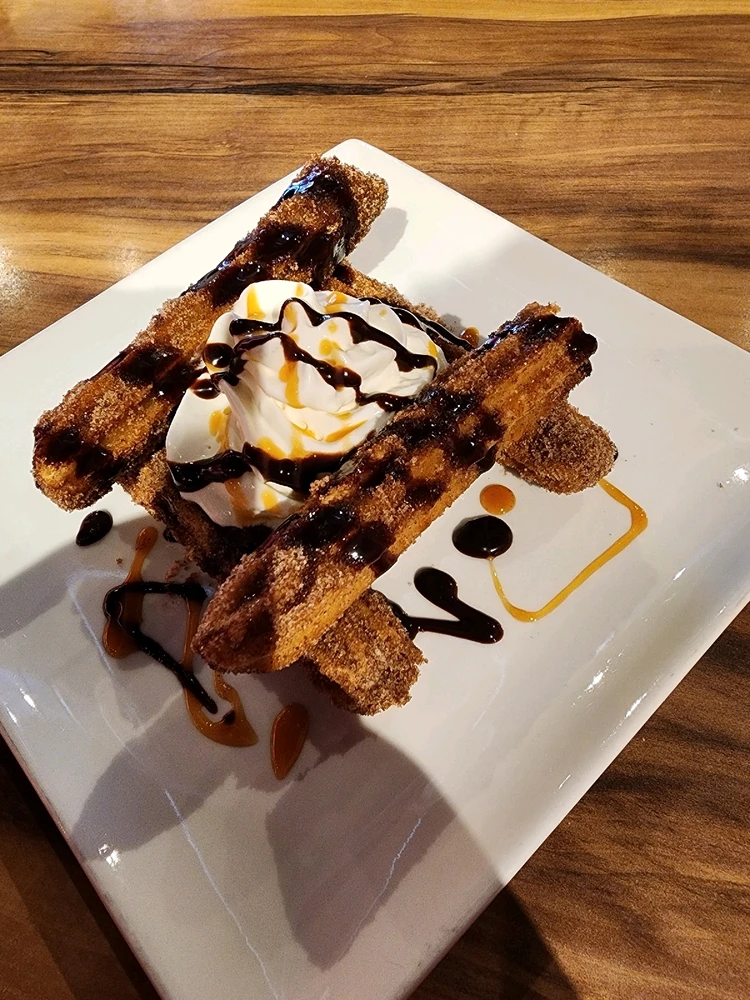 Churros with whipped cream, chocolate syrup, caramel syrup on a white square plate