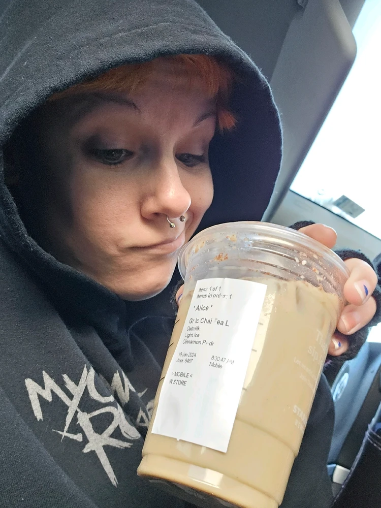 Woman wearing hoodie holding an iced chai looking longingly at the drink