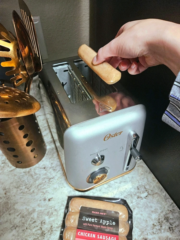 An open bag of chicken sausage on a counter next to a toaster, and a hand holding one sausage over a toaster slot.