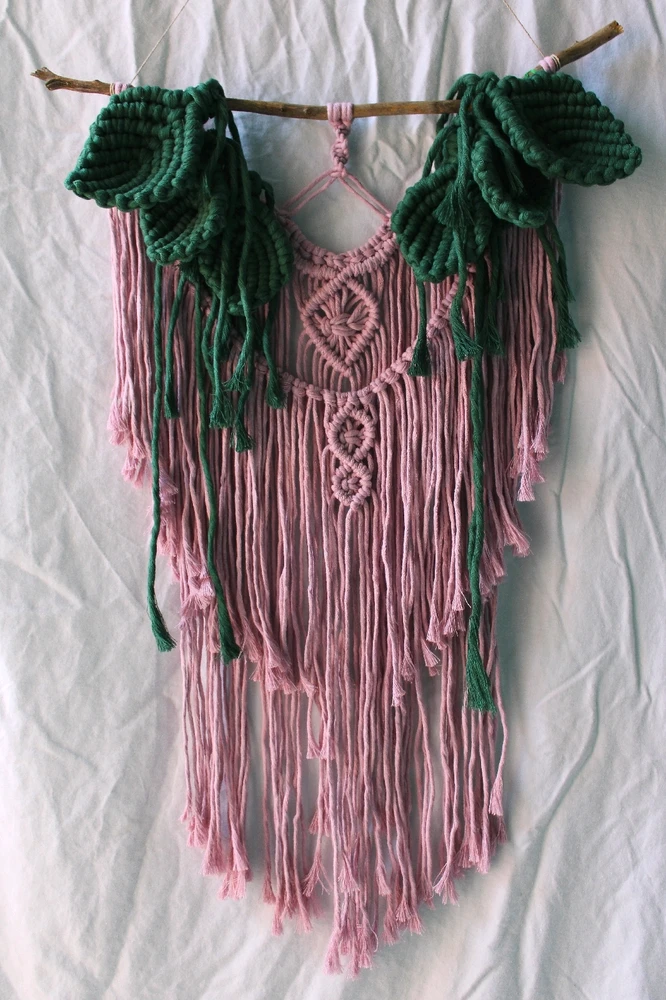 A pale pink boho style macrame piece featuring large green macrame leaves trailing inwards from the top edge.