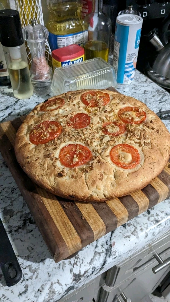 Round loaf of focaccia bread resting on a cutting board. The loaf is topped with tomato slices, garlic, and herbs.