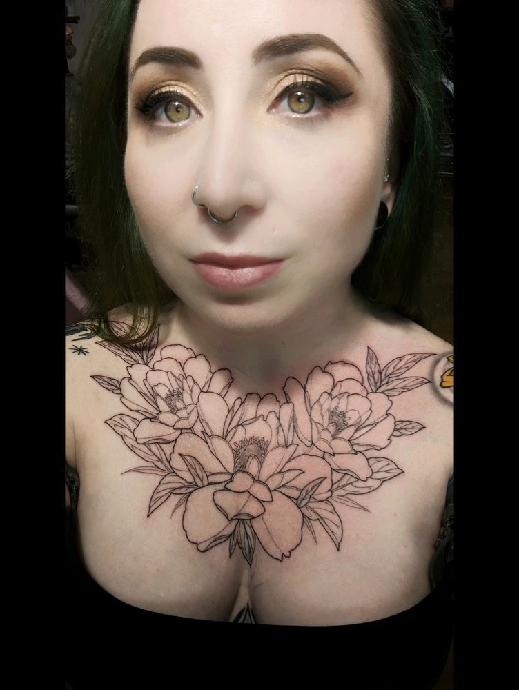 Girl with green hair and green eyes looks into camera. You can see freshly tattooed outlines of peonies on her chest.