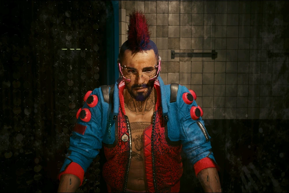 A rough looking man with sleeves rolled up, pink and blue jacket, and a red and blue mohawk. 