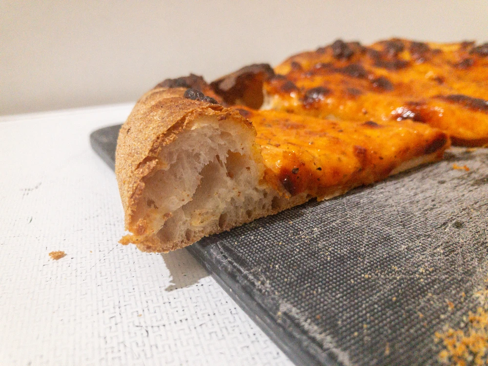 A side on shot of a slice of pizza, showing a tall crust with a bubbly interior as the centre of the shot.