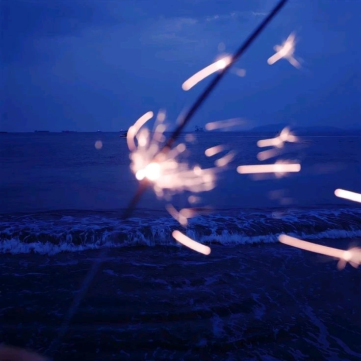 A person stands near a body of water, holding a sparkler that illuminates the night with a warm glow.
