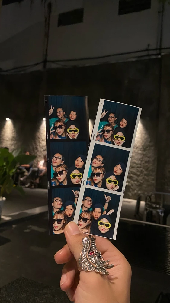 Holding 2 papers of 3-picture photo booth prints, each in black and white border. Photos of a group of not-close friends.