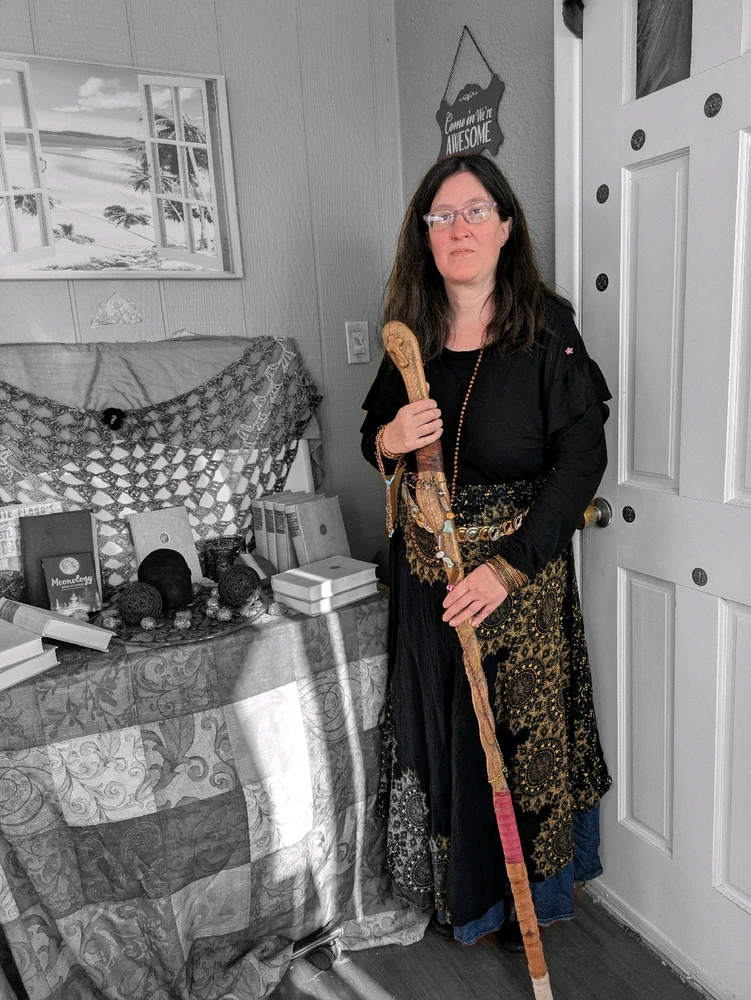 Woman dressed in witches garb, with a staff in hand, standing next to a witch's table decorated with candles, skulls, books 
