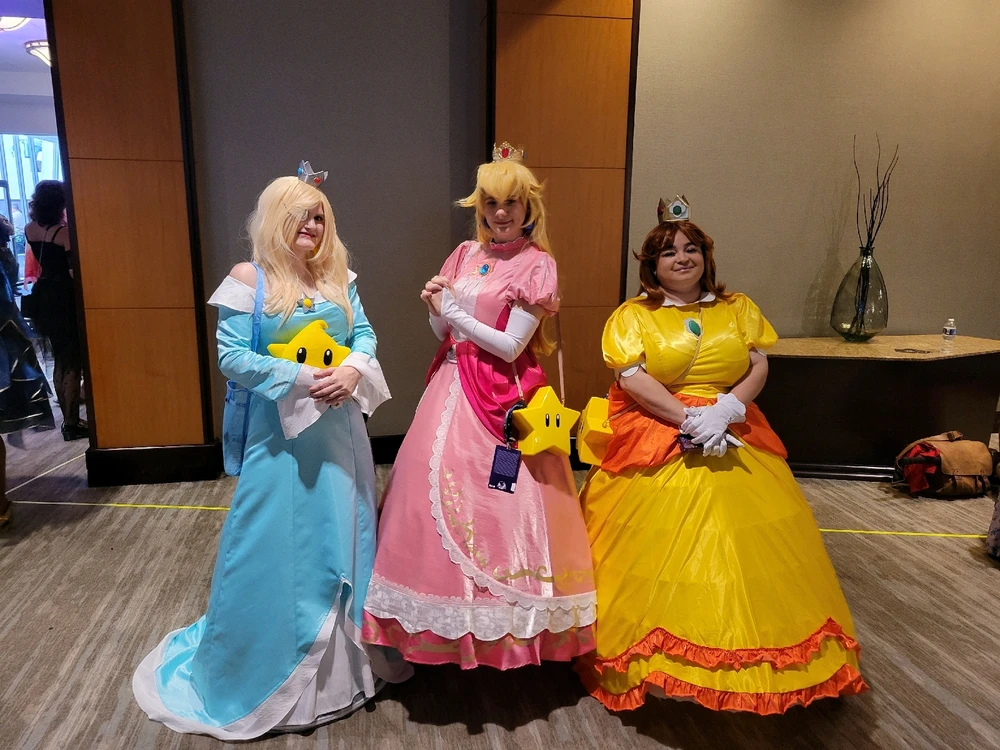 A cosplay picture of Rosalina, Peach and Daisy from Super Mario Bros. Each wearing a long dress of cyan, pink and yellow