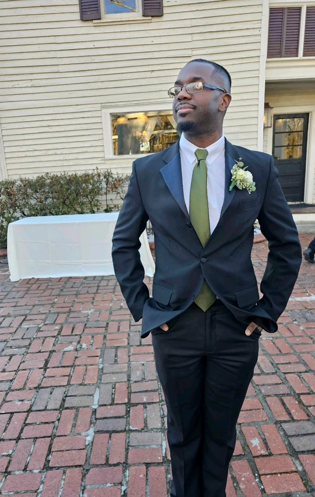 An extremely handsome man dressed in a black suit, with a white button down shirt and an olive green tie.