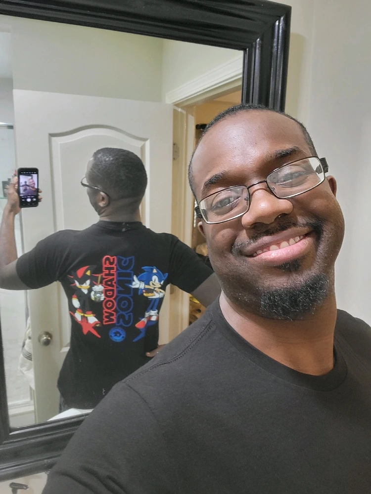 An incredibly handsome black man, sporting a black tee shirt with Sonic and Shadow the Hedgehog on his shirt.