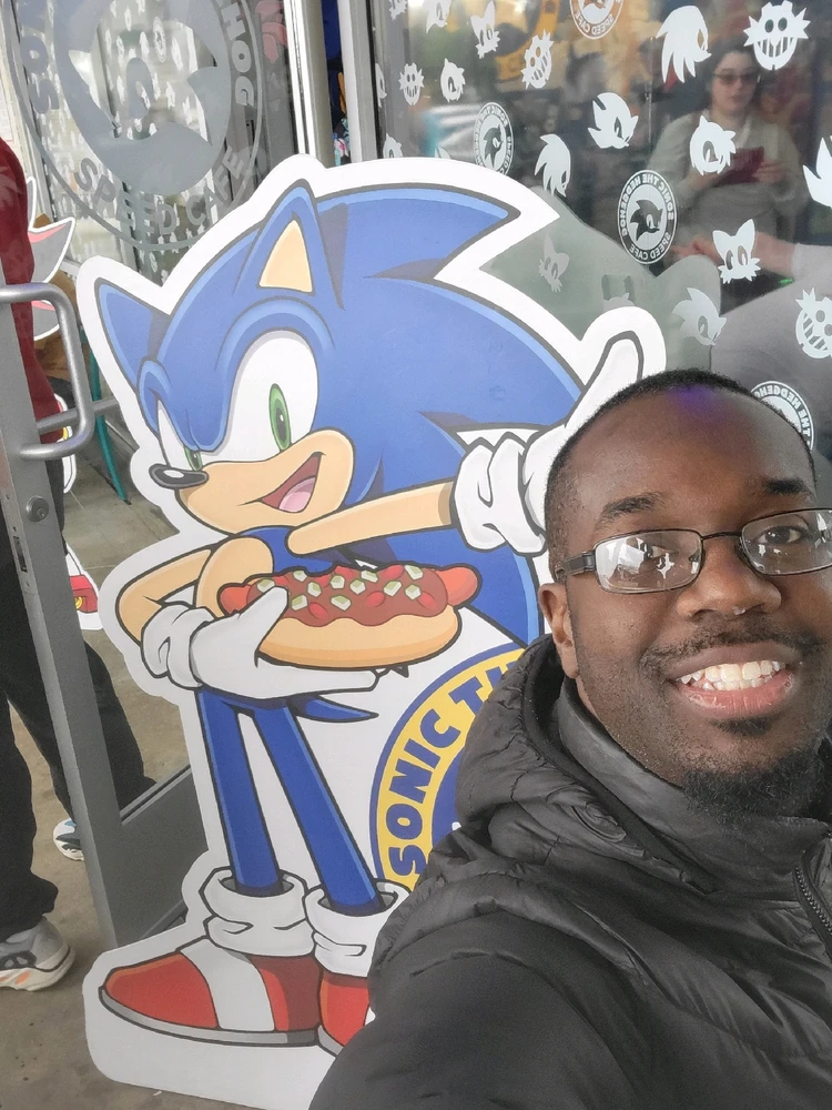 An incredibly handsome black man posing next to a Sonic cardboard outfit. He is smiling like a dork. 