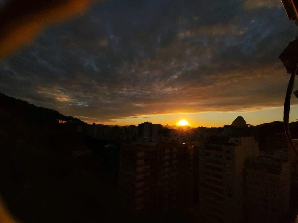 the sun peeks from behind the buildings and the mountain range, the golden rays bless the city. the sky is huge blue-ish grey
