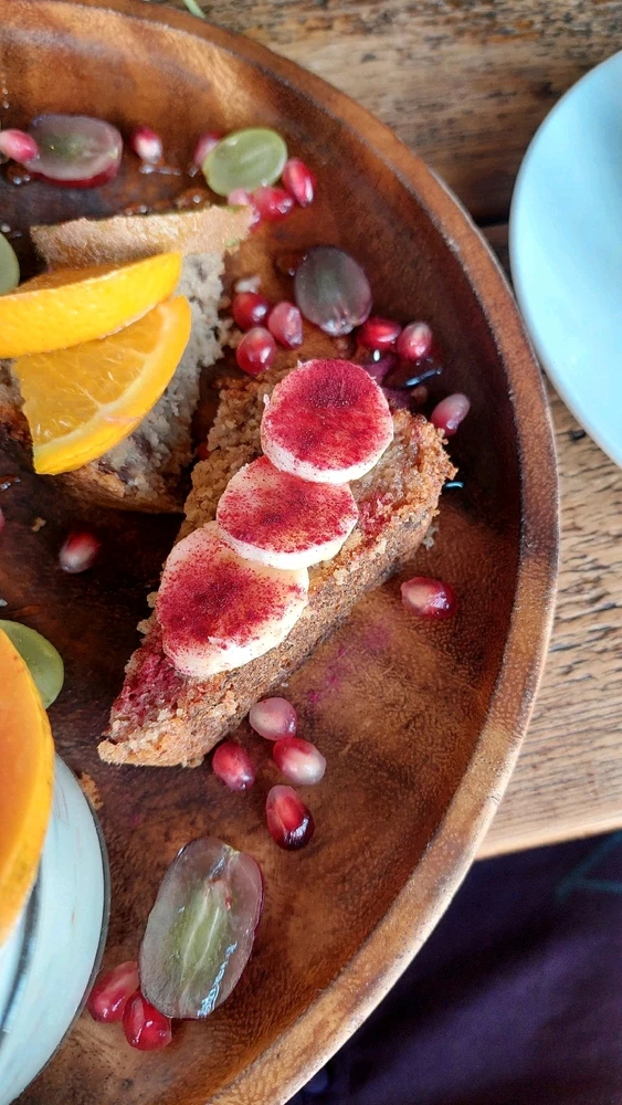 Bananenbrot mit Babanenstücken drauf und darüber Erdbeerpulver und auf dem anderen Bananenbrot frische Orangescheiben.