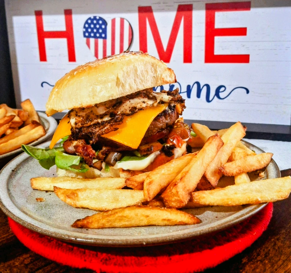 Cheese burger on honey butter sesame bun with bacon, tomatoes, relish, and onions and topped with a fried egg and fries.