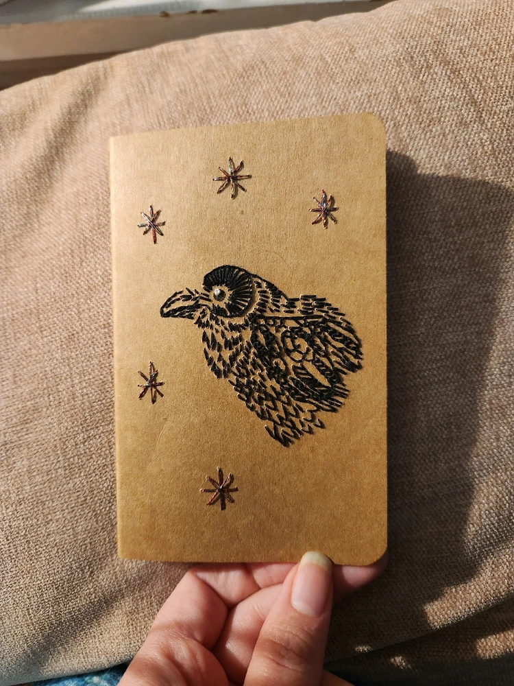 Small, brown paper notebook with an embroidered raven in the middle. There are 5 sparkly stars around the raven.