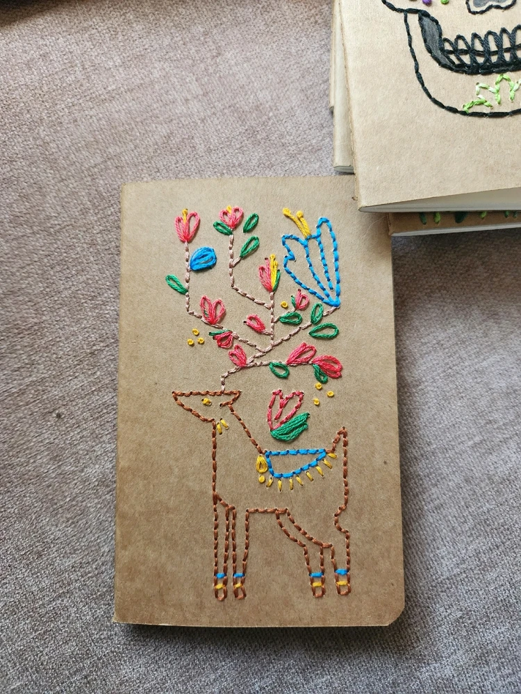 Small, brown paper notebook with a stylized deer embroidered on it. There are flowers coming out of the deer's antlers.
