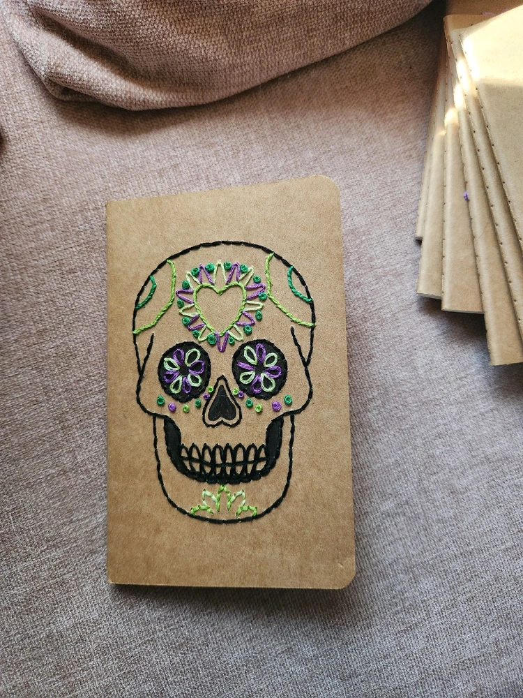 Small, brown paper notebook with a sugar skull embroidered into the cover. Main color theme is green and purple.