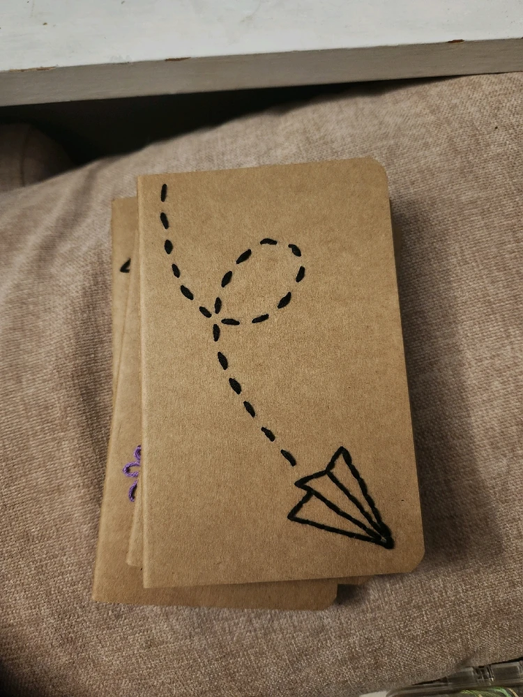 Small, brown paper notebook with a paper airplane embroidered onto the cover. The trail of the plane is visible.