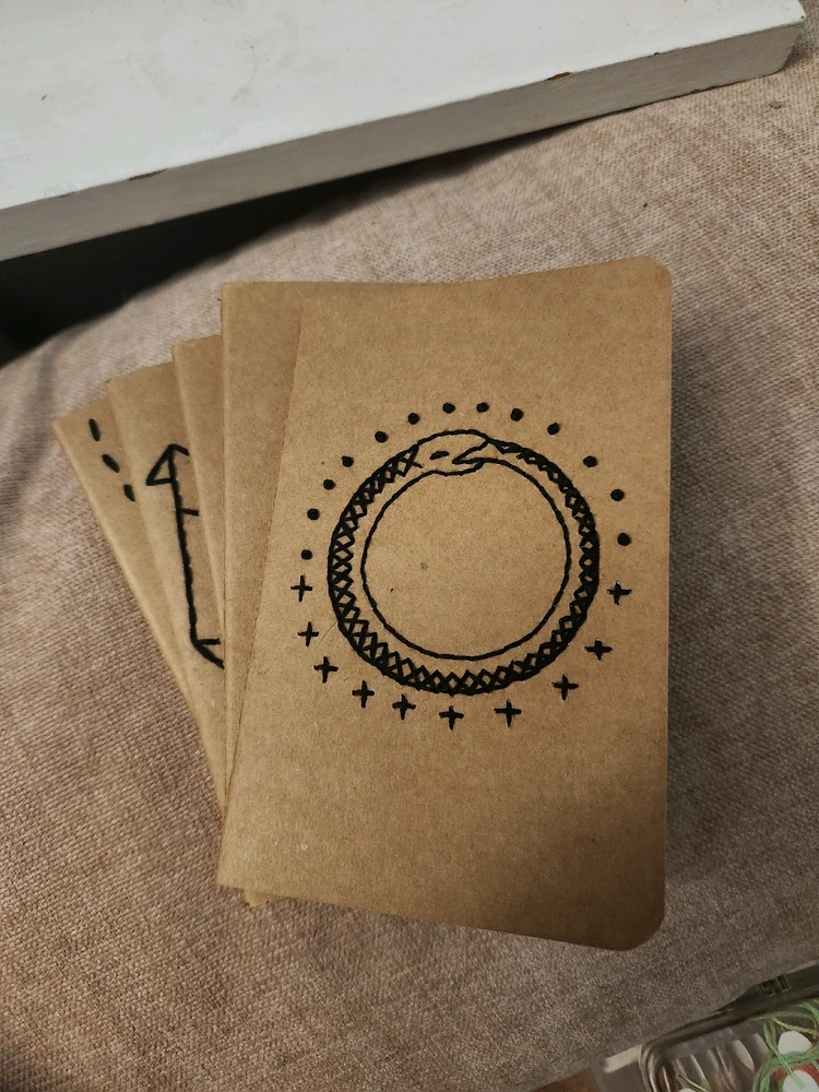 Small, brown paper notebook with an ourobouros embroidered on the cover. Stars surround the tail-eating serpent.