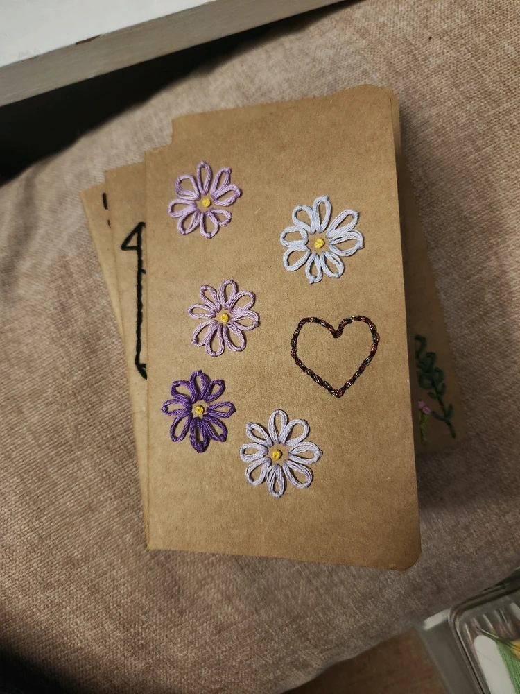 Small, brown paper notebook with flowers and a heart embroidered on the cover. Main color scheme is shades of purple.