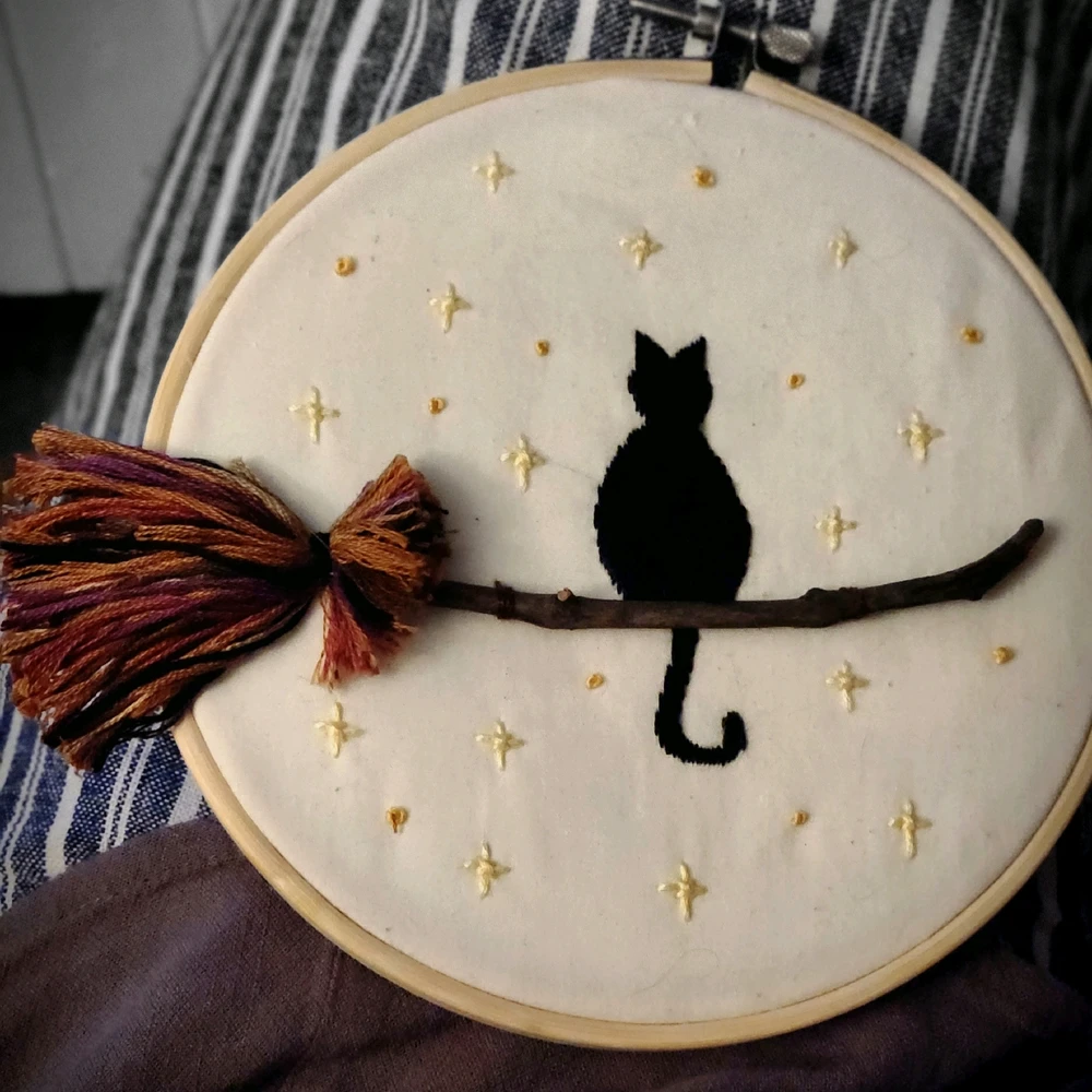 Witches broom is embroidered onto a hoop with a black cat sitting upon it. There are stars in the background.