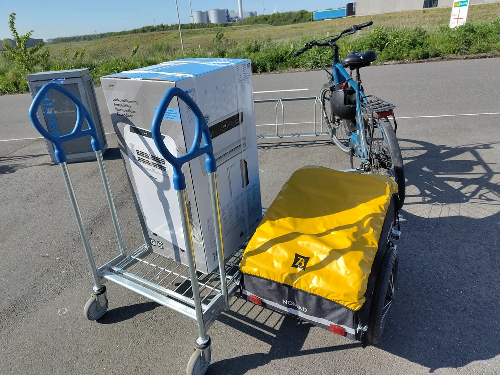 A box on a trolly beside a bike with a trailer attached to it. The package looks to be bigger than the trailer
