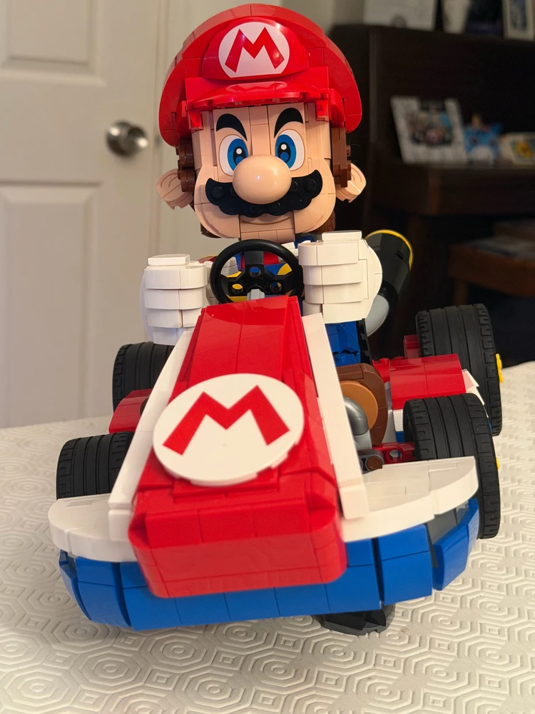 LEGO Mario sits in a colorful go-kart facing forward, with a red cap, blue eyes, and signature “M” logos displayed.