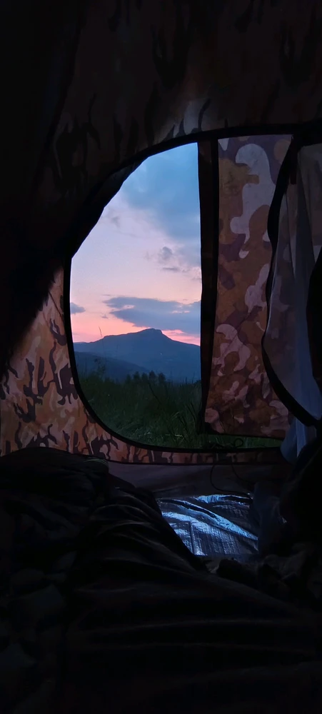 view from the dark inside of a camouflage camping tent looking out at a beautiful sunset over a mountain ridge in the distanc