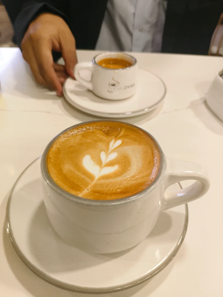 A close-up of a freshly brewed latte in a white cup with heart-shaped latte art, placed on a cafe table with another coffee 