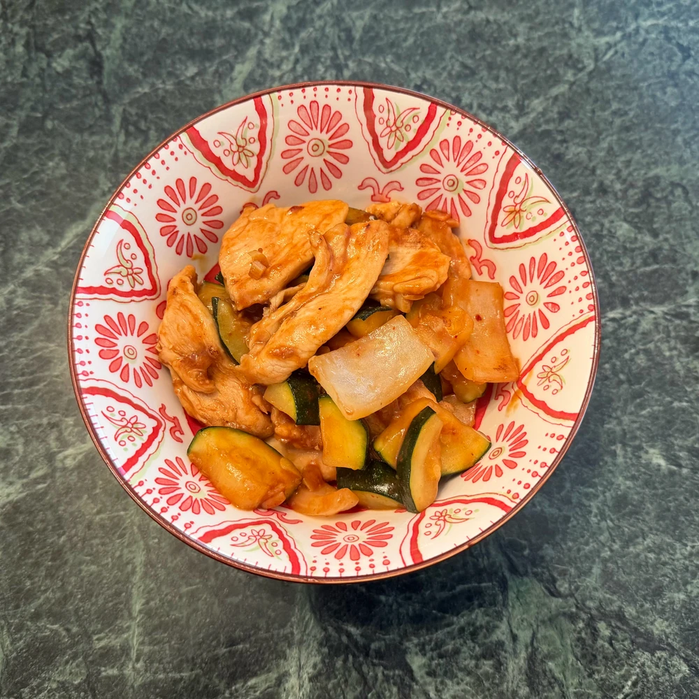 Red and white patterned bowl half full of stir fey garlic chicken with zucchini on a green marble tabletop 