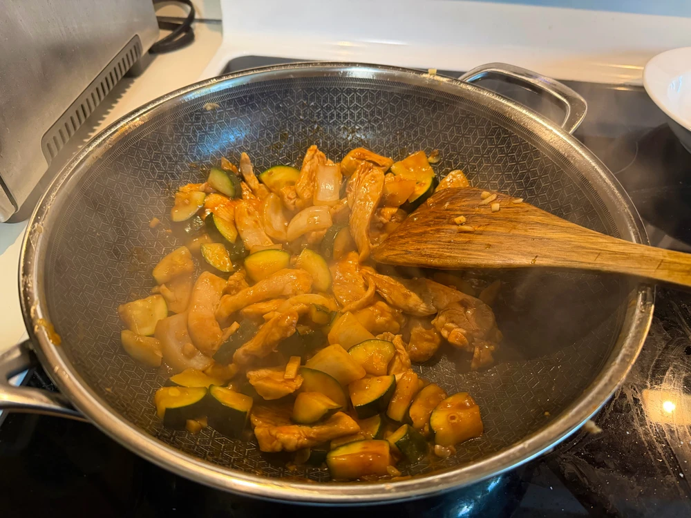 Wok filled with garlic chicken stir fry and zucchini with a wooden spatula in the wok on a glass top stove next to a toaster 