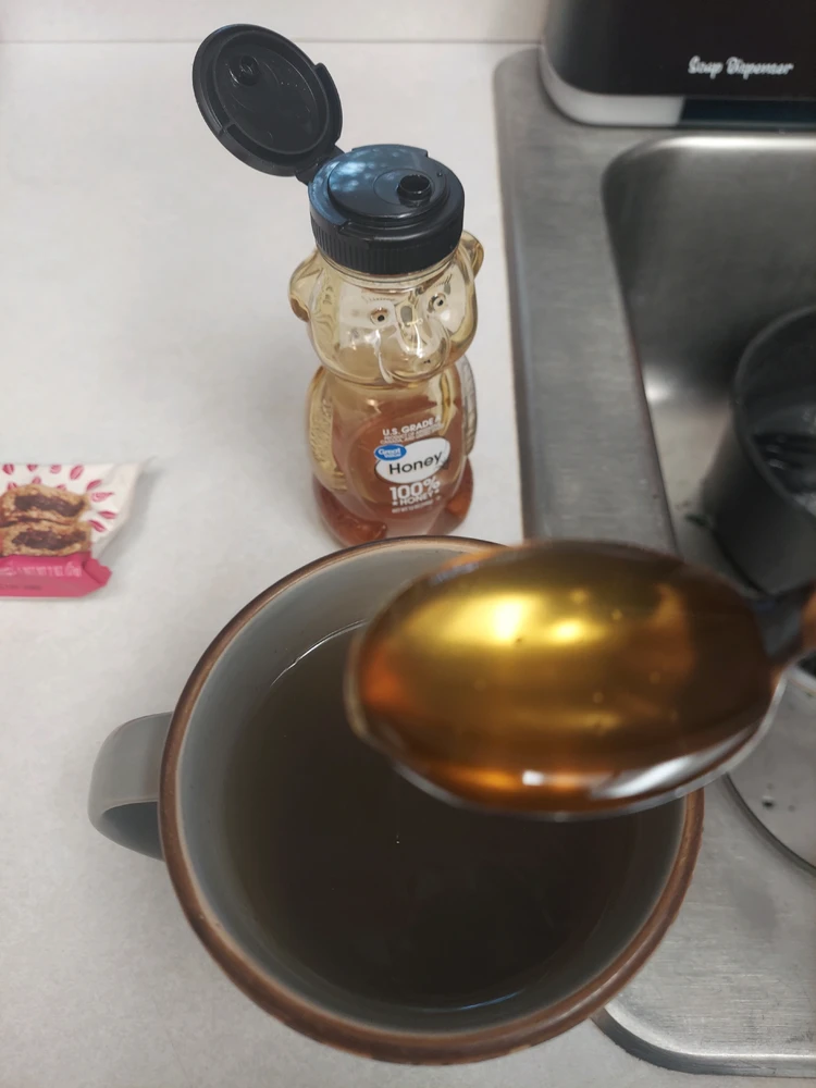 Spoonful of honey hovering over cup of tea. Honey container shaped like a bear in background next to a sink.
