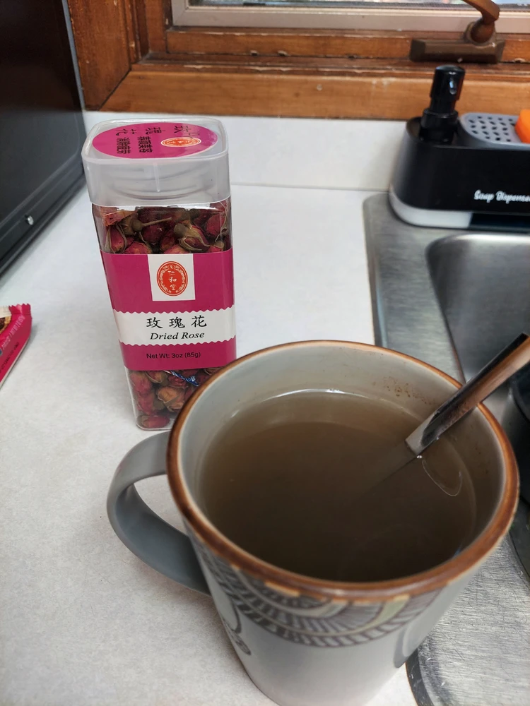 Container filled with rosebuds behind cup of tea with a spoon sitting in it next to a sink.
