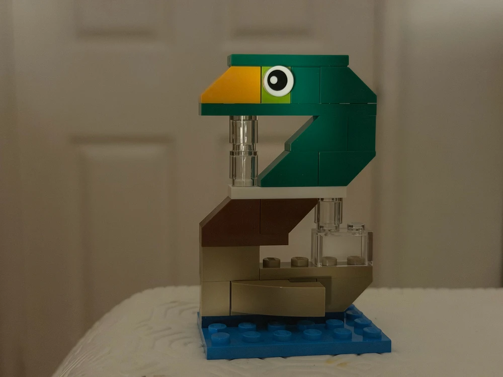Teal and tan LEGO duck with a yellow beak on a blue base, on a white textured surface indoors.