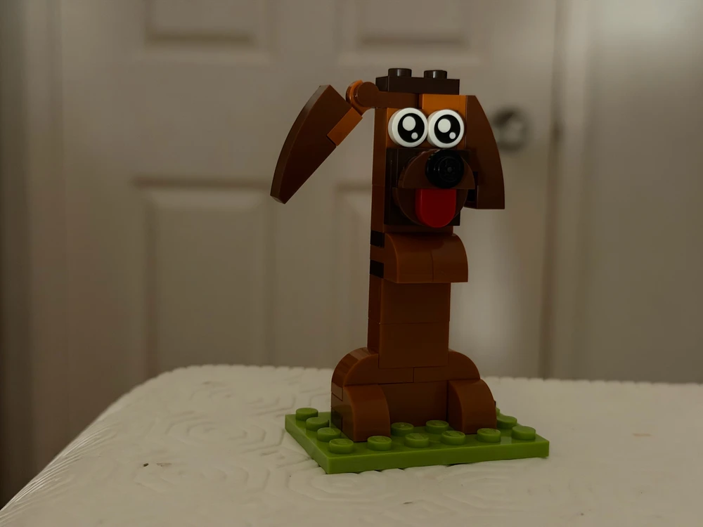 Brown LEGO dog with floppy ears and big eyes on a green base, on a white textured surface indoors.