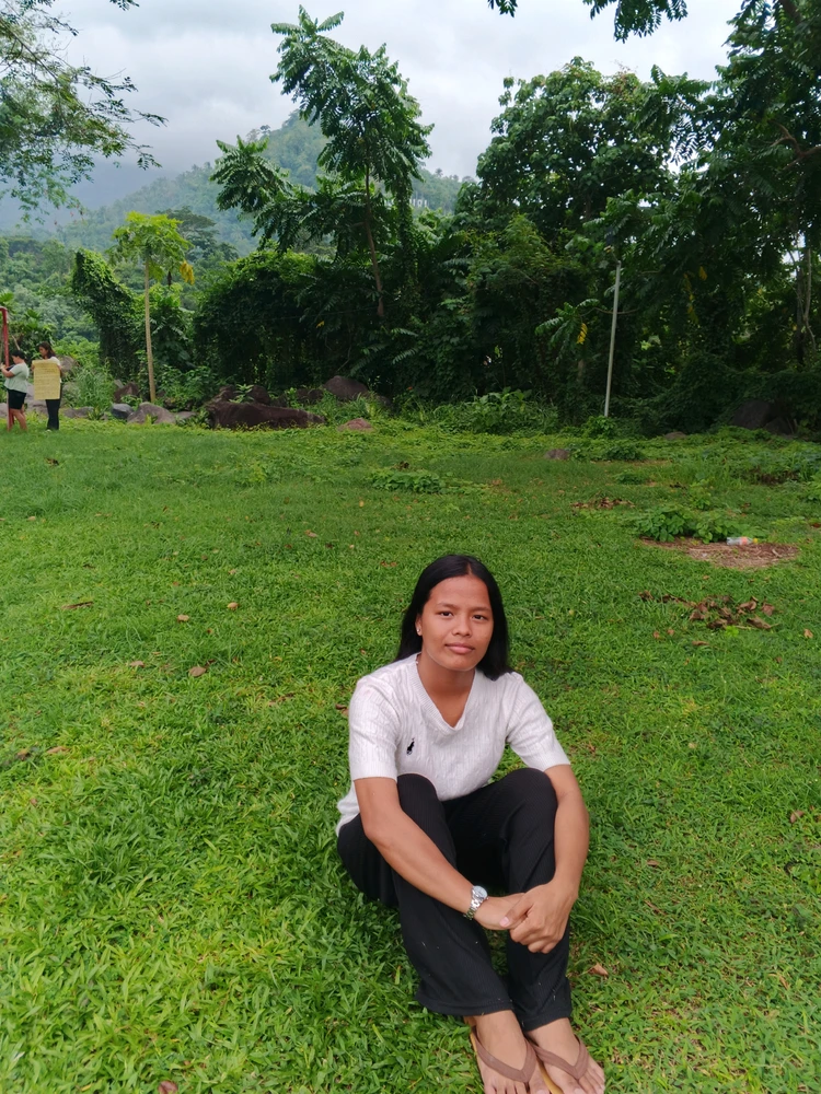 a person sitting on green grass outdoors, surrounded by trees and plants.