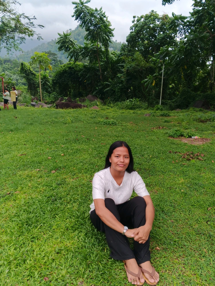 a person sitting on green grass outdoors, surrounded by trees and plants.