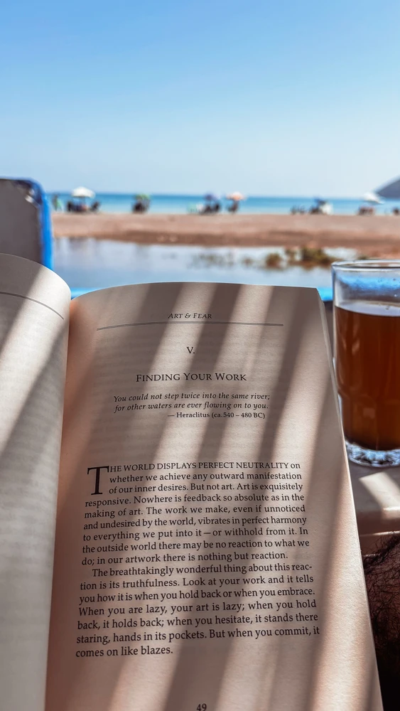 An open book featuring a Heraclitus quote, beside a glass of tea overlooking the sea.