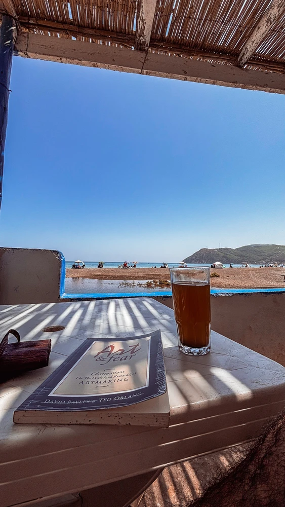 A book and glass of tea on a seaside table, with a quiet beach and mountain in the background.